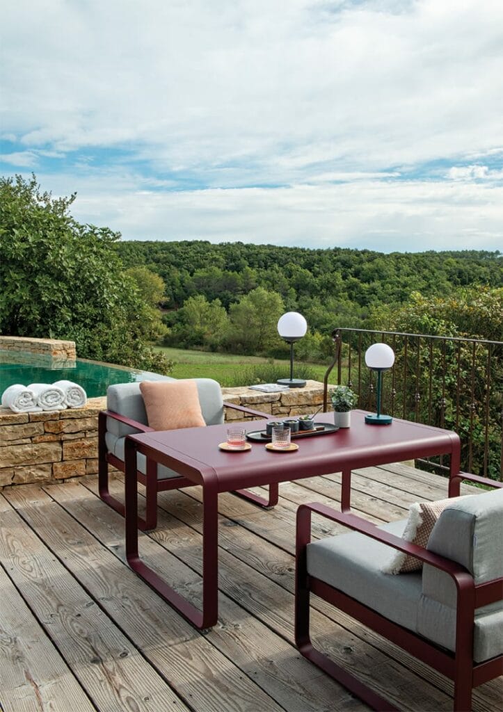 Patio extérieur avec une table rouge, des sièges confortables des Jardins du Faubourg et des collines verdoyantes pittoresques en arrière-plan, décoré dans une esthétique moderne et décontractée.
