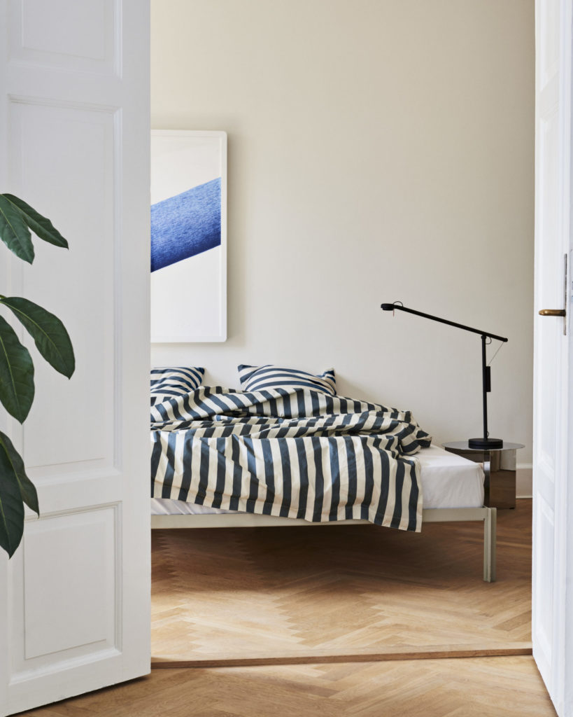 Une Lampe de bureau Fifty Fifty dans une pièce aux murs blancs et au doudou rayé bleu et blanc.