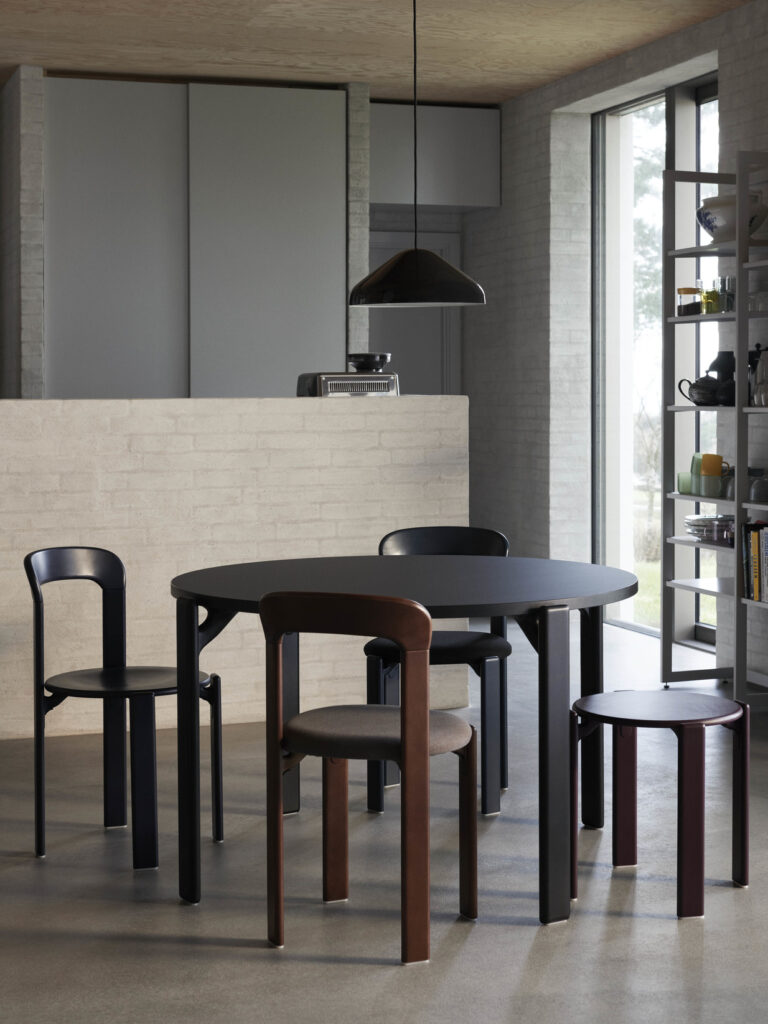 Dans un espace de restauration contemporain, la table Expo - Rey en noir occupe le devant de la scène, entourée de quatre chaises assorties, sur fond de mur de briques claires. Au-dessus, une suspension éclaire la scène et des étagères remplies de divers accessoires ajoutent de la profondeur à l'arrière-plan.