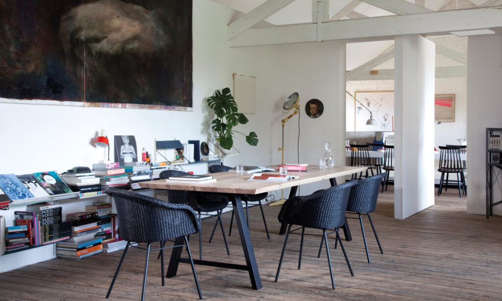 Une pièce moderne est dotée d'une table Albert en bois, de chaises noires, de livres et d'œuvres d'art sur les murs. Une grande plante trône dans le coin, complétée par le parquet en bois et les poutres blanches apparentes.