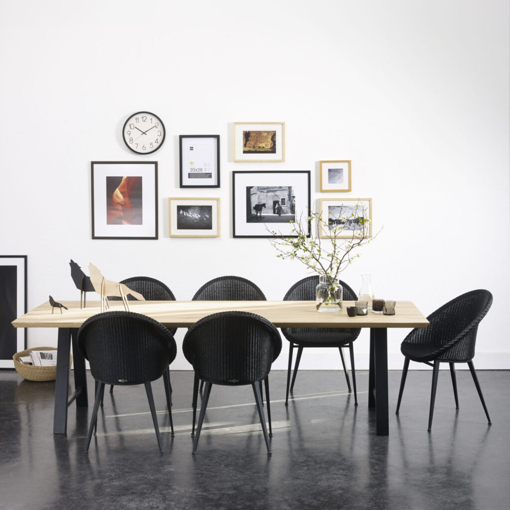Une salle à manger présente une table Albert en bois avec six chaises noires autour, des œuvres d'art et une horloge sur le mur, et un vase avec des branches sur le dessus de la table.