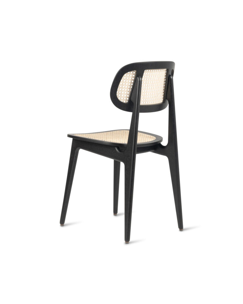 La Chaise Titus est une chaise moderne avec une structure en bois noir, un dossier et une assise en rotin tressé, avec des pieds fuselés et un design minimaliste.