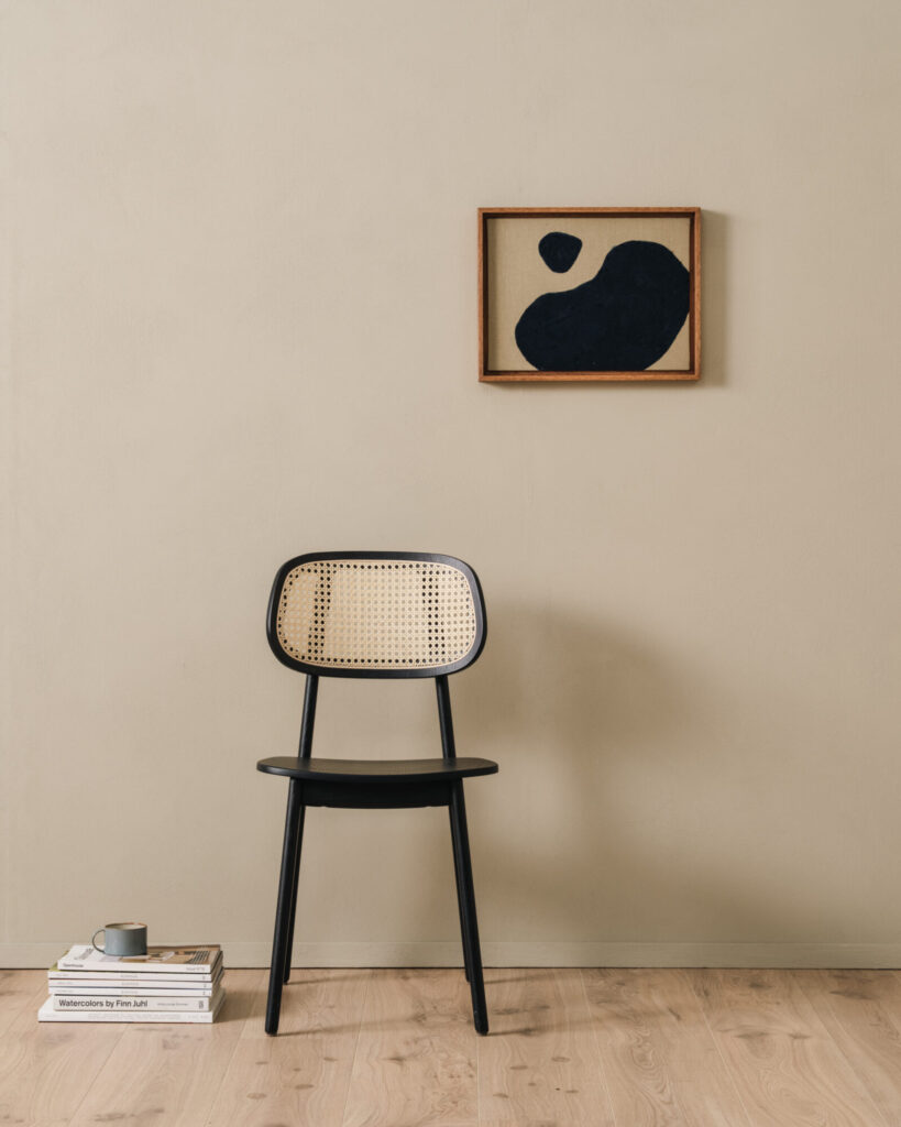 Contre un mur beige, la chaise Titus en bois au dossier tressé trône sous un tableau abstrait noir et blanc. Une pile de livres et un mug à proximité renforcent l'atmosphère invitante à la détente.