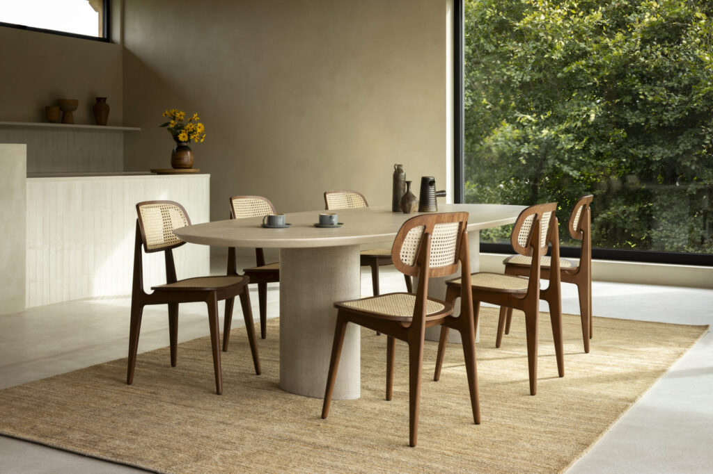 Une salle à manger moderne est dotée d'une table ronde avec six chaises en bois Chaise Titus. La table est ornée de petites tasses noires et une grande fenêtre offre une vue sur la verdure luxuriante à l'extérieur.