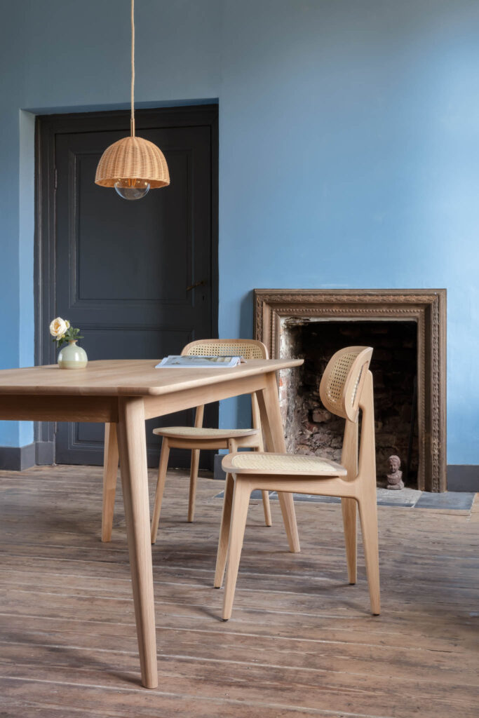 La pièce bleu clair est dotée d'une table à manger en bois avec des chaises Chaise Titus, complétée par une cheminée et une suspension. Un petit vase avec une fleur sur la table ajoute du charme.