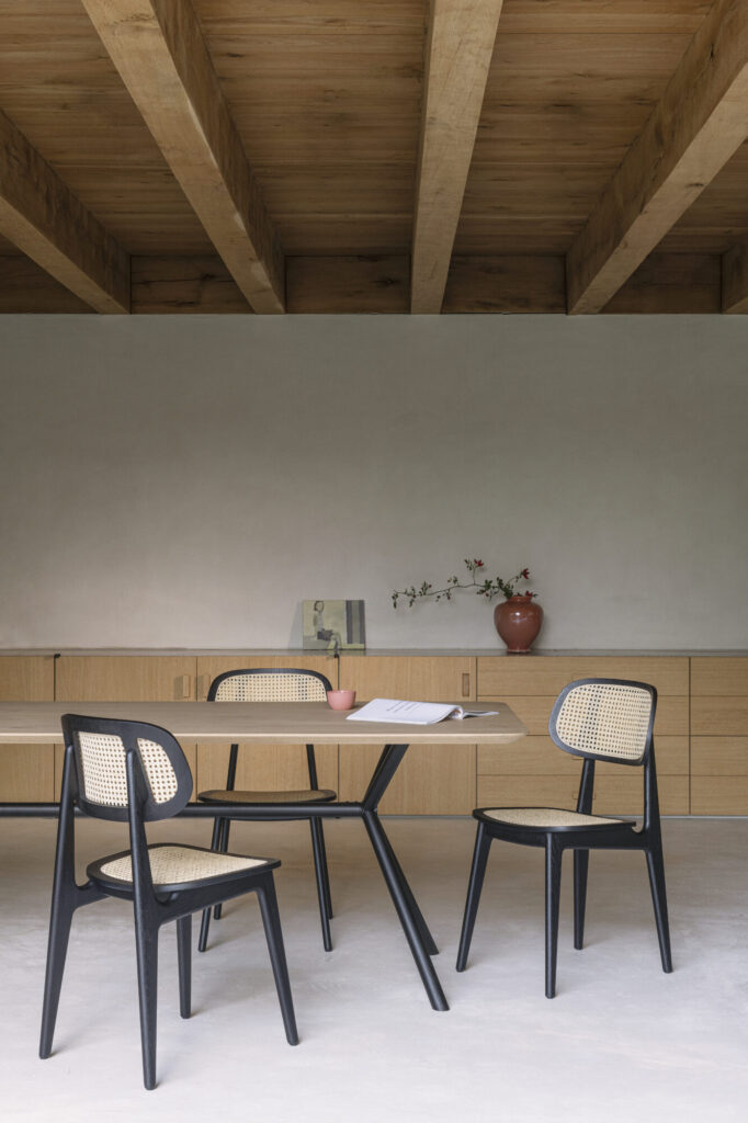 La salle à manger met en valeur une table en bois entourée de quatre chaises Chaise Titus, avec des cadres noirs et des dossiers tressés. Un vase de fleurs et un livre ornent la table, complétée par un meuble latéral et des poutres en bois apparentes.