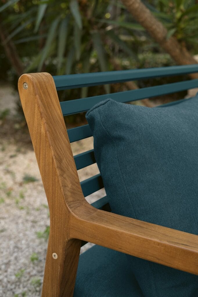 Gros plan d'une chaise de luxe Fauteuil Somerset en bois et tissu bleu sarcelle, avec un coussin moelleux, sur un fond de jardin flou.