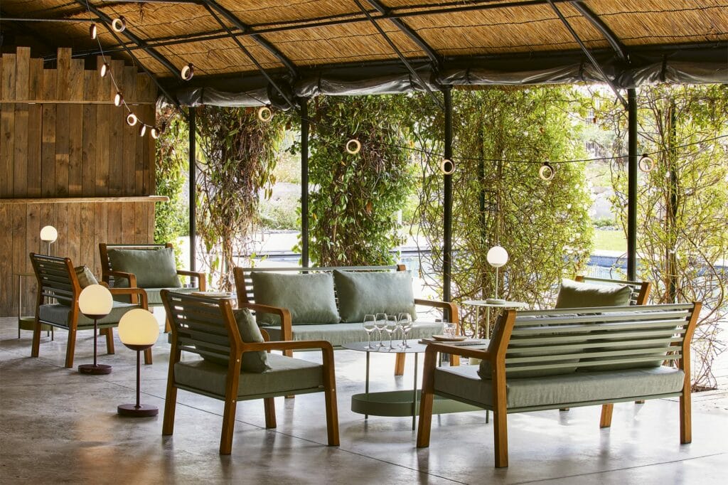 Coin salon extérieur confortable avec canapés verts et fauteuil Somerset, murs en bois et plantes suspendues, sous un toit de chaume près d'un plan d'eau.