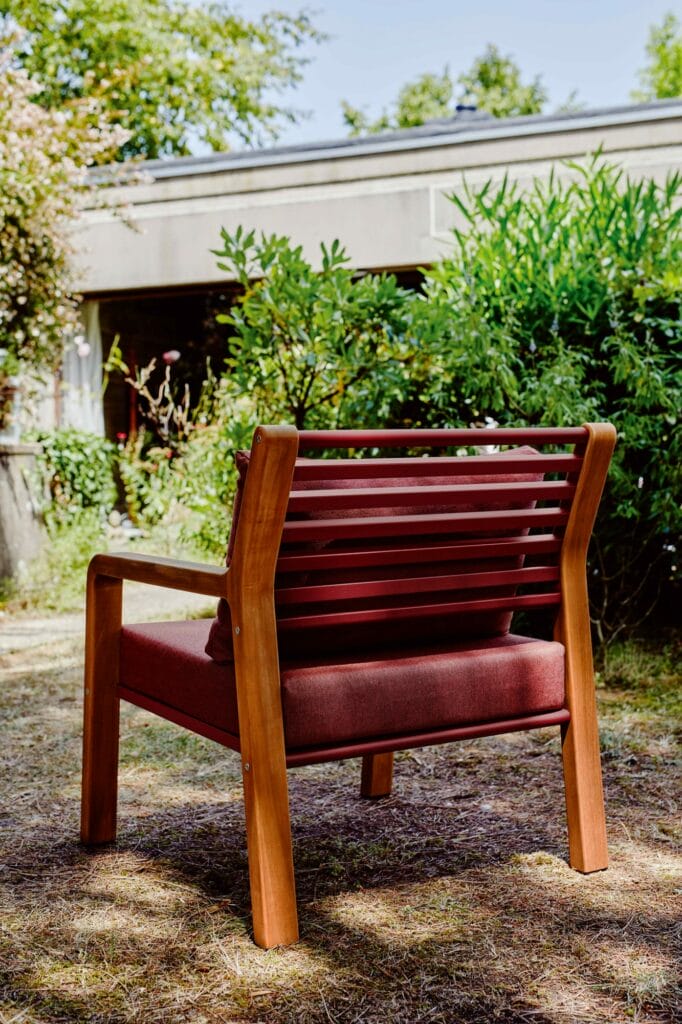 Une chaise Fauteuil Somerset moderne avec des coussins rouges posés sur l'herbe dans un jardin ensoleillé, avec une verdure luxuriante et une structure en béton en arrière-plan.