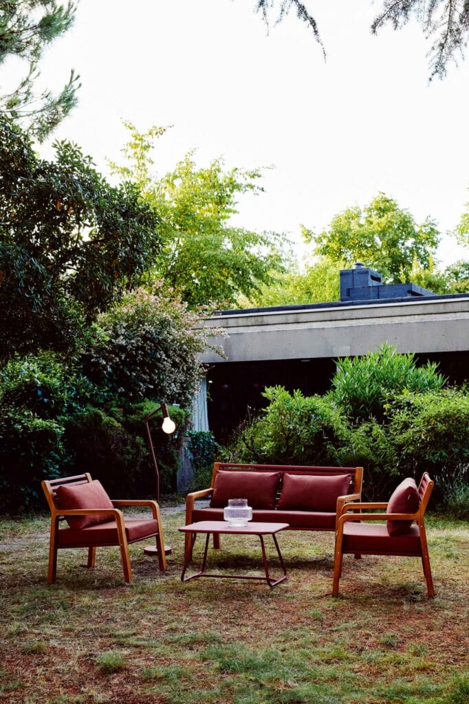 Un ensemble de sièges d'extérieur confortable comprenant une chaise Fauteuil Somerset marron, deux fauteuils assortis et une petite table avec une lampe, posés sur l'herbe contre une verdure luxuriante et une maison en arrière-plan.