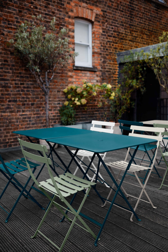 Une scène extérieure accueillante présente une Chaise Bistro en métal bleu sarcelle, entourée de chaises en métal colorées et d'une chaise longue confortable sur la terrasse en bois, avec des murs en briques et des plantes luxuriantes en toile de fond.