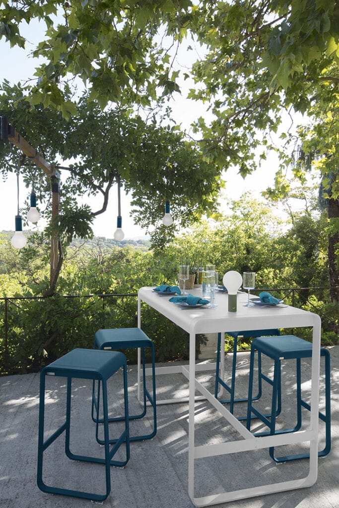 Une Table haute Bellevie, 140 x 80 cm et des chaises posées sur une terrasse avec des arbres en fond.