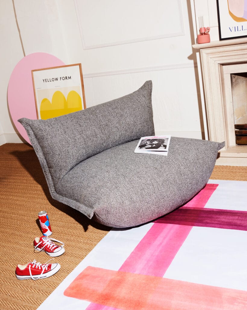 Un pouf gris est posé sur un tapis, les baskets BonBaron Mingle placées à côté.