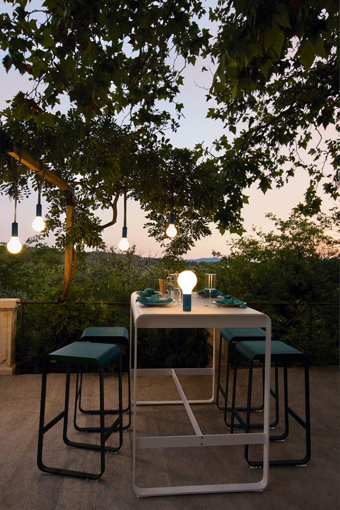 Une Table haute Bellevie, 140 x 80 cm et des chaises sur une terrasse sous un arbre.