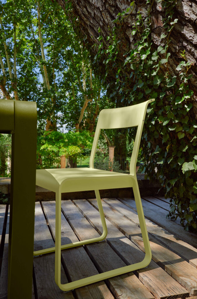 Une Chaise V2 Bellevie en métal vert clair est posée sur une terrasse en bois à côté d'une table, entourée de verdure et de troncs d'arbres recouverts de lierre.
