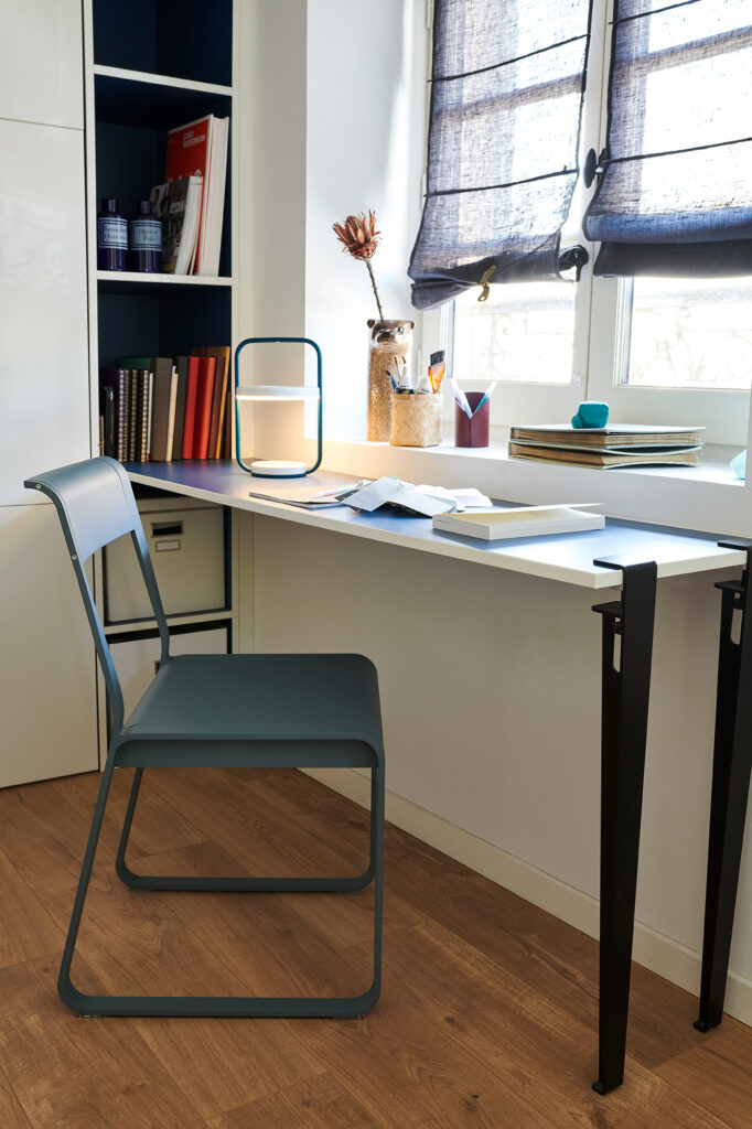 Un bureau moderne avec des livres, des papiers, une lampe et des décorations se trouve près d'une fenêtre avec des stores transparents ; la Chaise V2 Bellevie est placée devant le bureau.