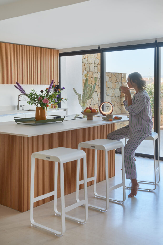 Assise en pyjama sur un tabouret de bar Bellevie, une tasse à la main, devant un îlot de cuisine moderne, une personne est assise. À travers de grandes fenêtres, la lumière du soleil filtre sur les fleurs et les fruits disposés sur le comptoir.