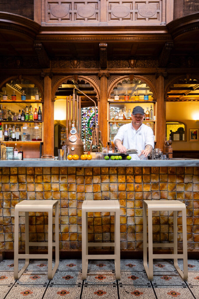 Un barman prépare des boissons derrière le bar, entouré de bouteilles et de fruits, tandis que trois tabourets de bar Bellevie sont positionnés avec style au comptoir.