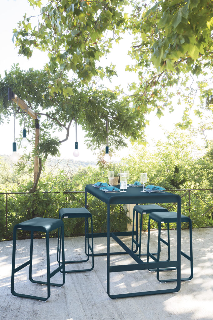 Un ensemble de salle à manger de patio contemporain comprend une table assortie et quatre tabourets de bar Bellevie en bleu sarcelle, disposés sous des arbres feuillus avec des plats et des verres posés dessus.