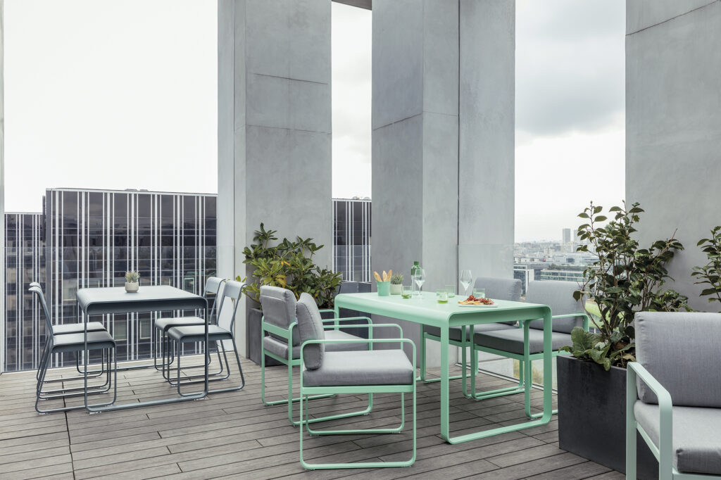 Une terrasse extérieure moderne présente la Table Bellevie (140 x 80 cm) en vert menthe et gris, entourée de plantes en pot, avec un horizon de la ville et un ciel nuageux en arrière-plan.