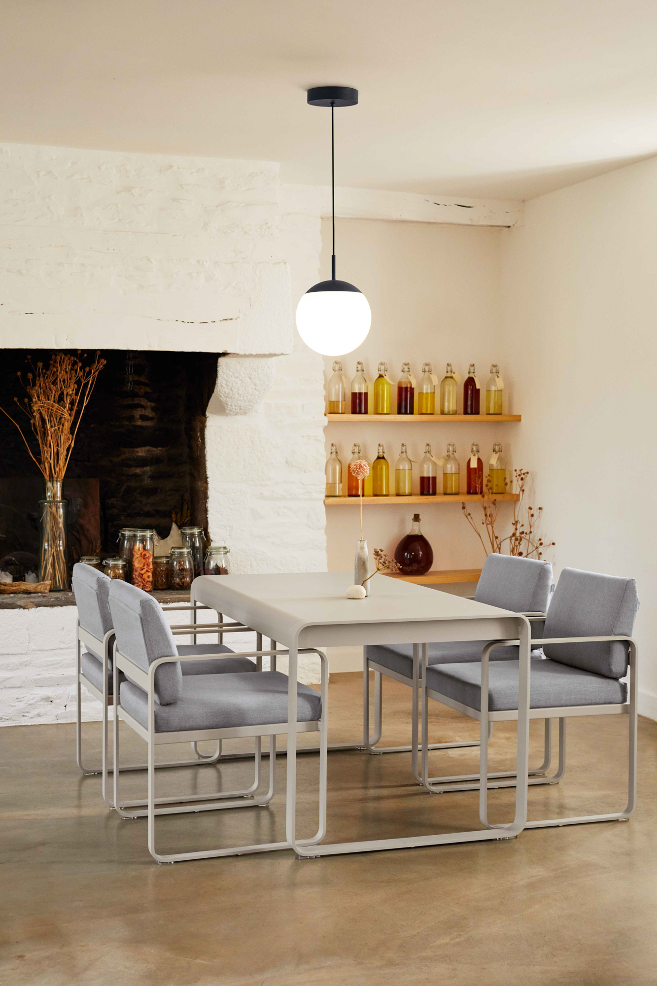 Une salle à manger minimaliste avec la table Bellevie, 140 x 80 cm, comme pièce maîtresse, entourée de quatre chaises grises rembourrées, avec des étagères de bouteilles en verre et une suspension moderne au-dessus.
