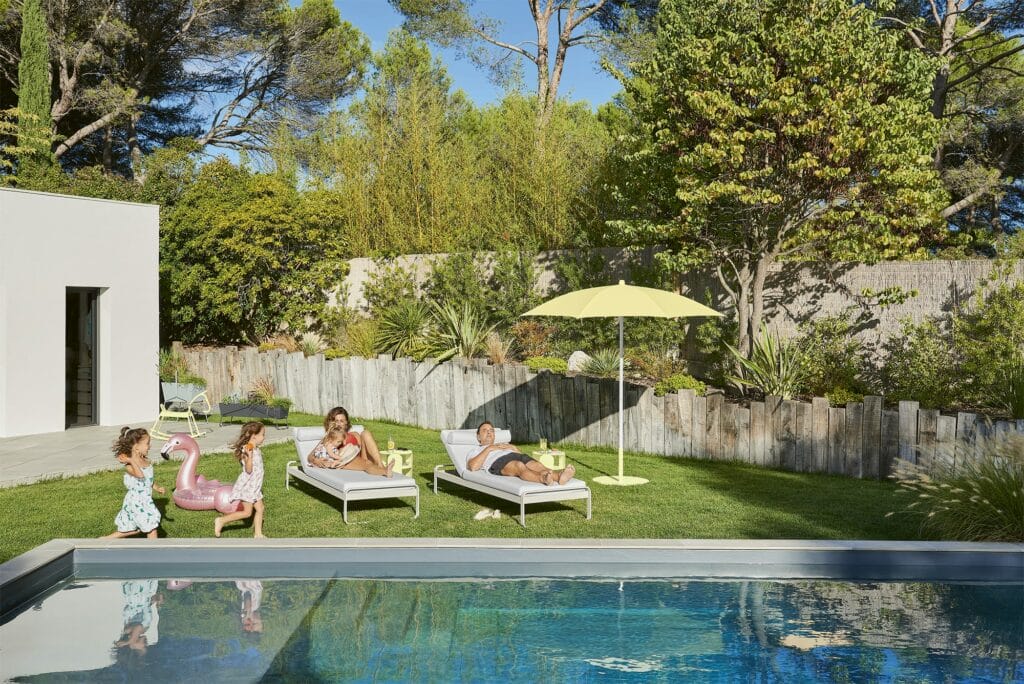 Famille profitant d'une journée ensoleillée au bord de la piscine dans une cour avec chaise longue Bellevie, parasol vert et arbres luxuriants en arrière-plan.