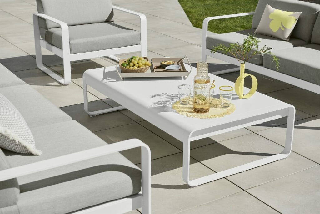 Phrase avec le nom du produit remplacé : Patio extérieur avec un mobilier blanc moderne, comprenant des chaises et une grande table basse Bellevie, orné de collations et d'un pichet de limonade sous le soleil.