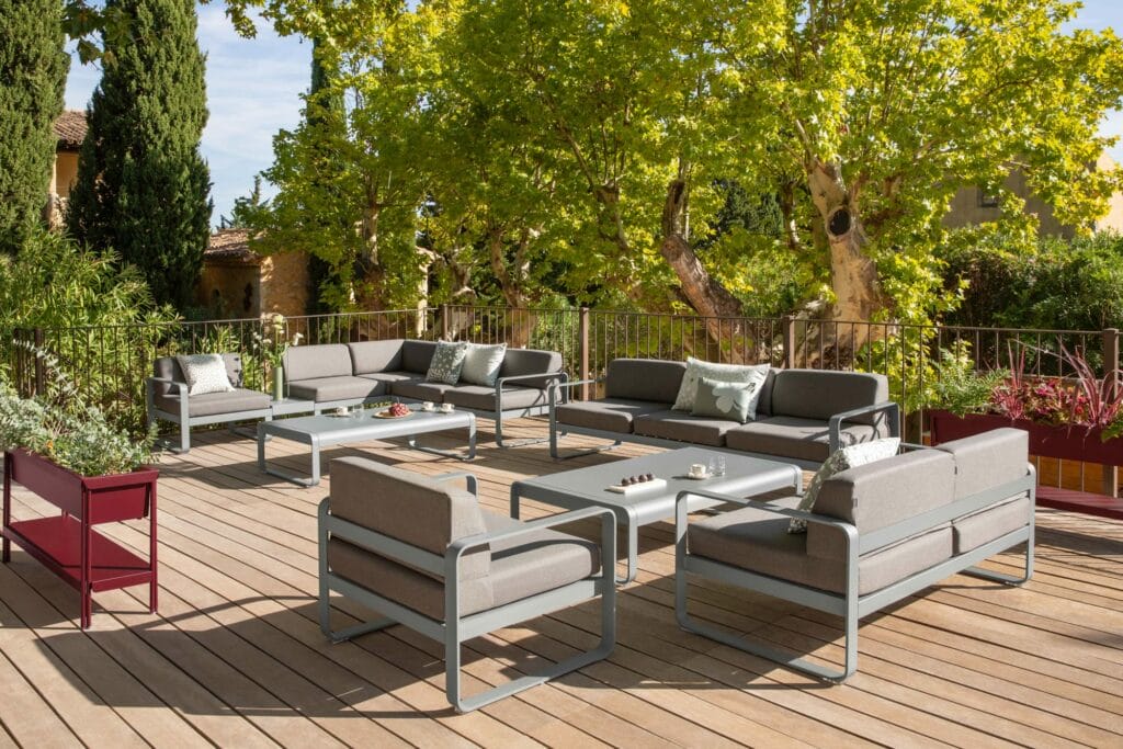 Patio extérieur avec mobilier moderne comprenant des canapés et une "Grande table basse Bellevie", installé sur une terrasse en bois entourée d'arbres et de plantes luxuriantes sous un ciel dégagé.