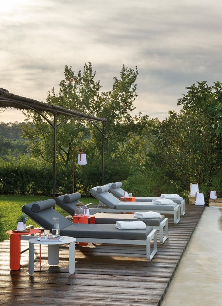 Un espace de détente extérieur serein avec plusieurs chaises longues Bellevies, tables d'appoint et lanternes, donnant sur une verdure luxuriante au crépuscule.
