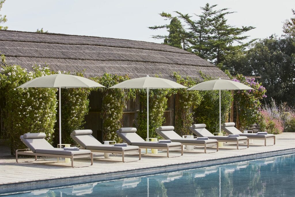 Cinq chaises longues Bellevie au bord de la piscine avec coussins beiges sous des parasols verts au bord d'une piscine, à côté d'un arbuste à fleurs et d'une cabane en bois au toit de chaume.