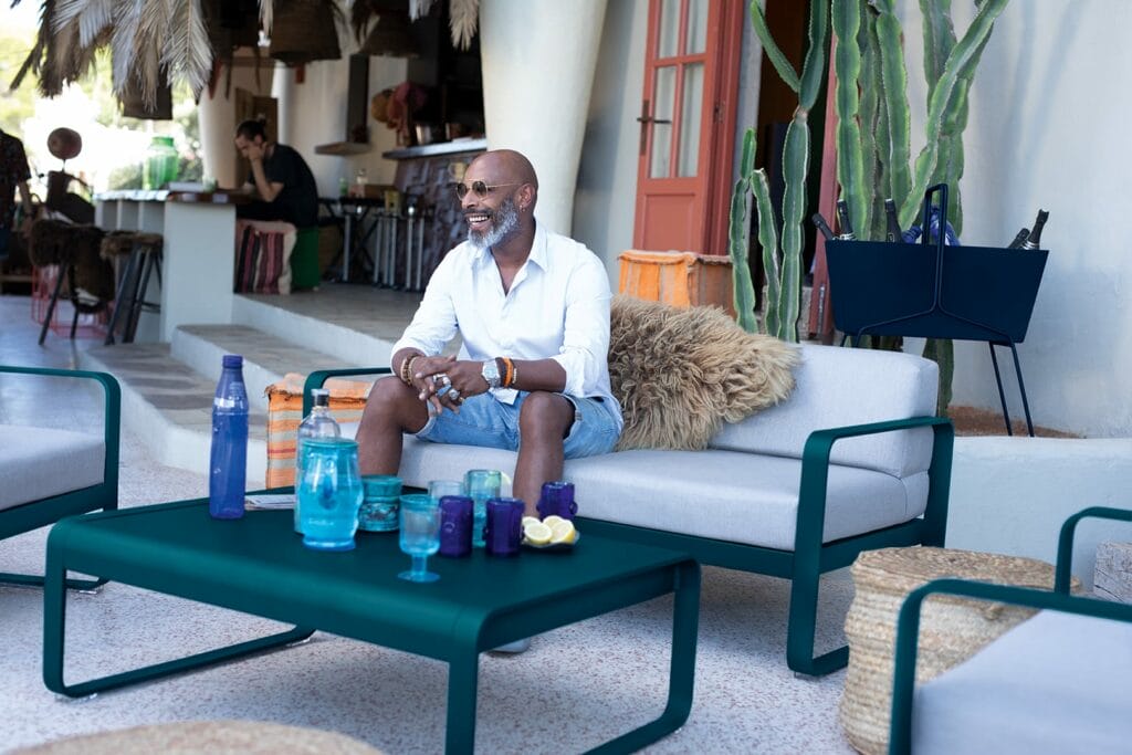 Phrase avec nom du produit : Un joyeux homme chauve assis sur un Canapé Bellevie 2 places, vêtu d'une chemise blanche et d'un short, tenant un verre d'eau, entouré de meubles de style et de plantes.