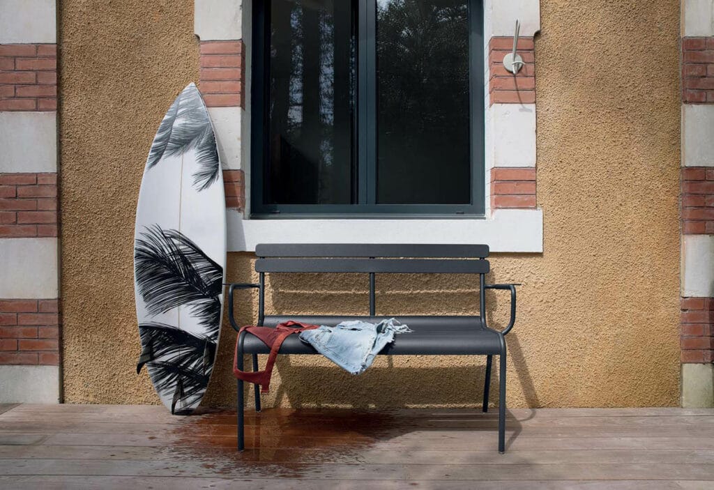 Un Banc Luxembourg avec accoudoirs devant une fenêtre, mettant en valeur une planche de surf.