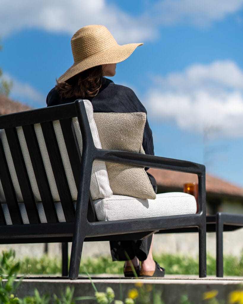 Une femme assise sur un banc, vêtue d'un Canapé de jardin Jack en teck noir - 2 places.