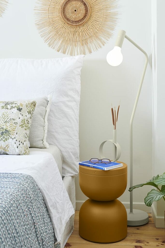 Un coin chambre confortable comprenant un lit blanc, une œuvre d'art murale décorative, une élégante table d'appoint Tabouret Piapolo avec des livres et un lampadaire.