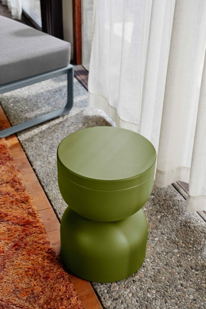 Un tabouret Piapolo cylindrique vert avec un couvercle, placé sur un tapis orange texturé à côté de rideaux blancs transparents et d'un cadre de lit gris.