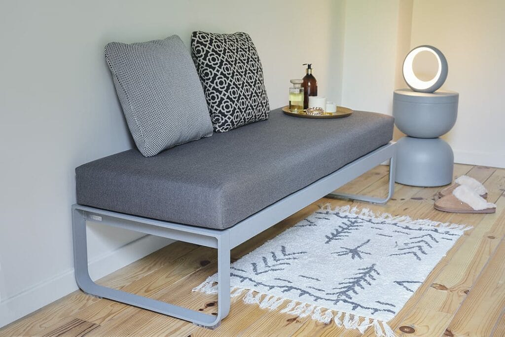 Lit de repos moderne et minimaliste avec revêtement gris et oreillers à côté d'une table d'appoint Lylee Round avec une lampe, sur un tapis à motifs dans une pièce lumineuse.