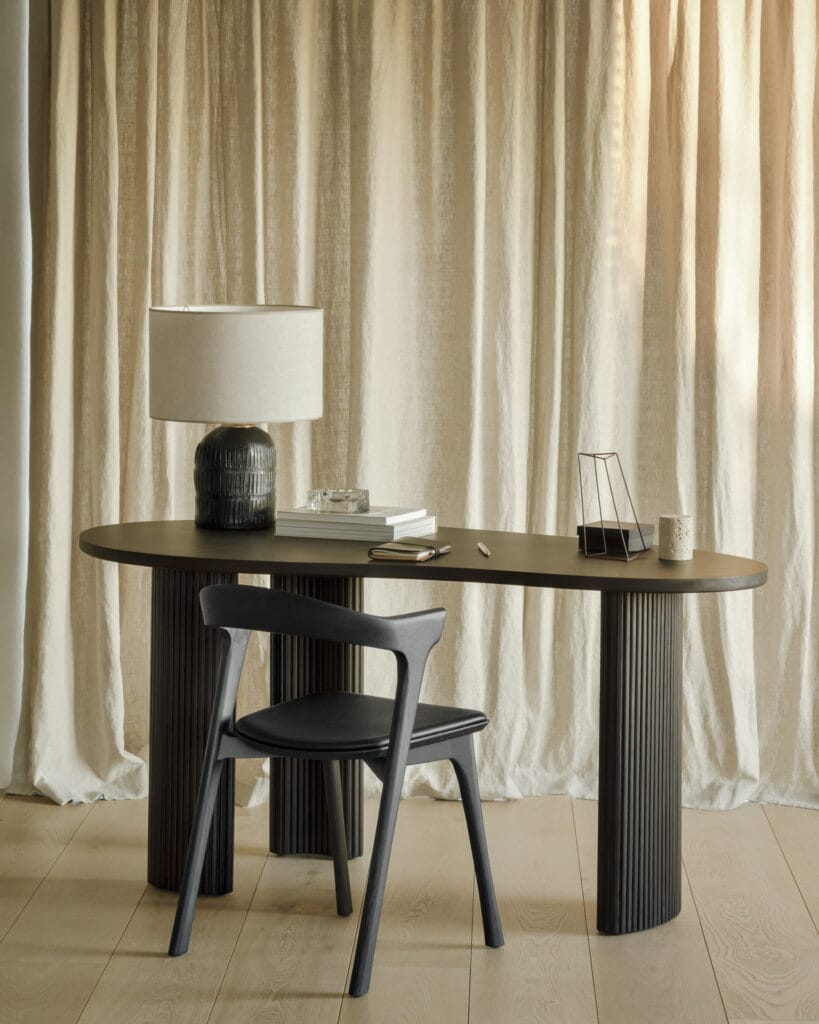 Un Bureau Boomerang en acajou avec une lampe et une chaise devant un rideau.