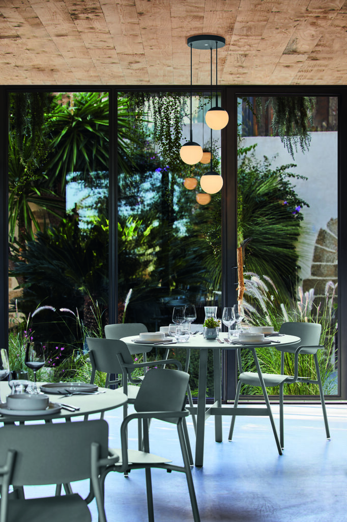 Un espace repas moderne avec des chaises vertes entoure la Table So'O, diam 117cm, placée près de grandes fenêtres donnant sur un jardin luxuriant avec de hautes plantes et une verdure vibrante.