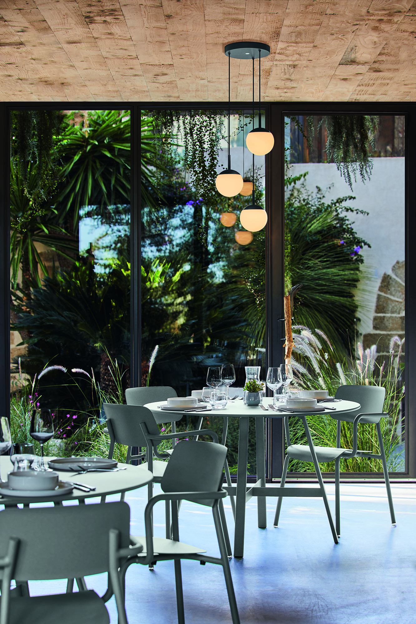 Un espace repas moderne avec des chaises vertes entoure la Table So'O, diam 117cm, placée près de grandes fenêtres donnant sur un jardin luxuriant avec de hautes plantes et une verdure vibrante.