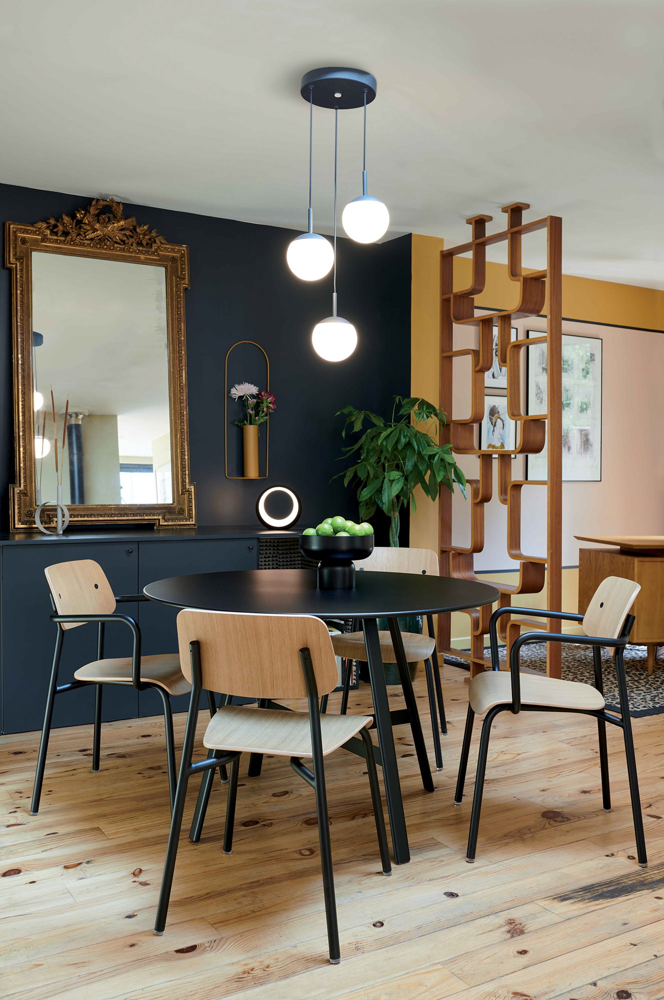 Une salle à manger moderne avec l'élégante table So'O (diam 117cm), quatre chaises en bois, des suspensions, un miroir à cadre doré, un séparateur de pièce en bois et un parquet en bois clair.