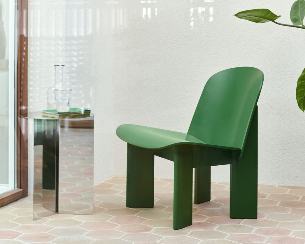 La chaise longue Chisel, dotée d'un revêtement vert moderne et de lignes courbes, est placée sur un sol carrelé à côté d'une table d'appoint réfléchissante avec des verres et des livres, près d'une fenêtre et d'une plante en pot.