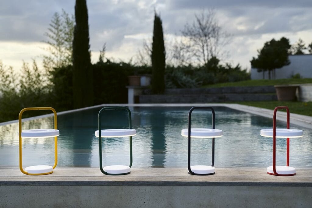 Quatre lampes LED portables Ulli Lampe H30 avec poignées colorées alignées sur un rebord au bord de la piscine au crépuscule, surplombant une piscine sereine et un jardin paysager.
