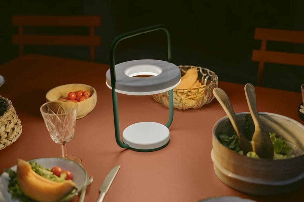 Un Philips Hue Go moderne et portable illumine une table à manger dressée avec divers plats, notamment des bols de salade et de fruits, ainsi qu'un verre d'eau.