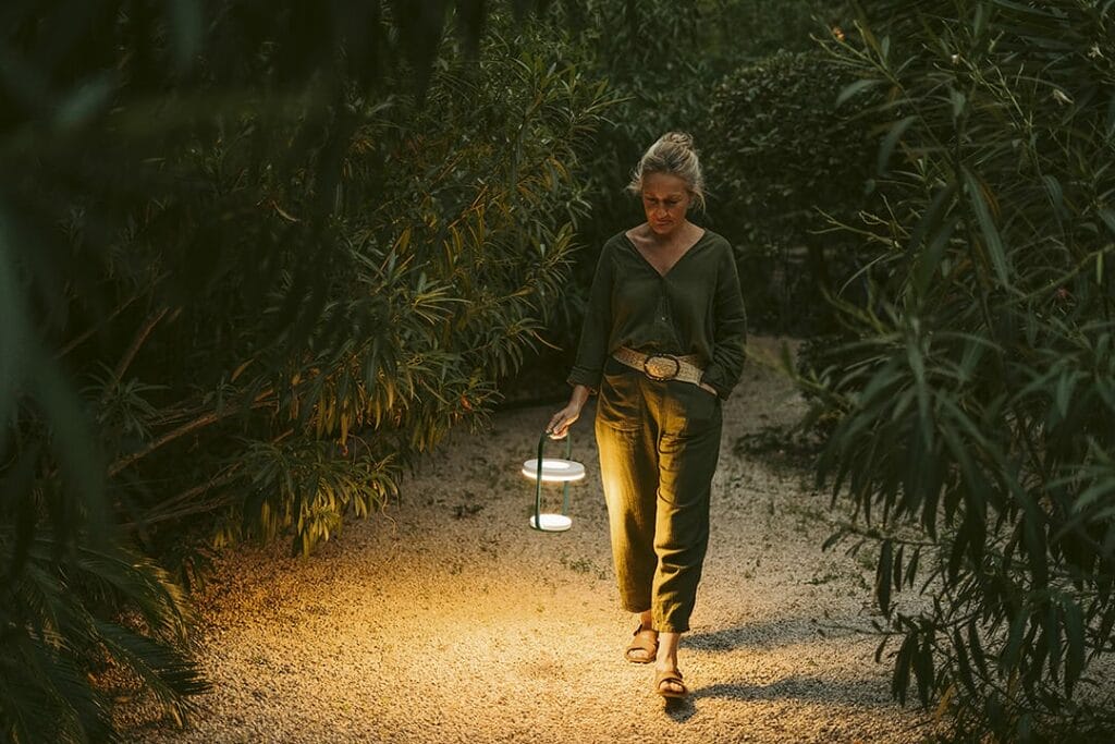 Une femme marche le long d’une allée de jardin au crépuscule, tenant une Ulli Lampe H30 qui éclaire le sol.