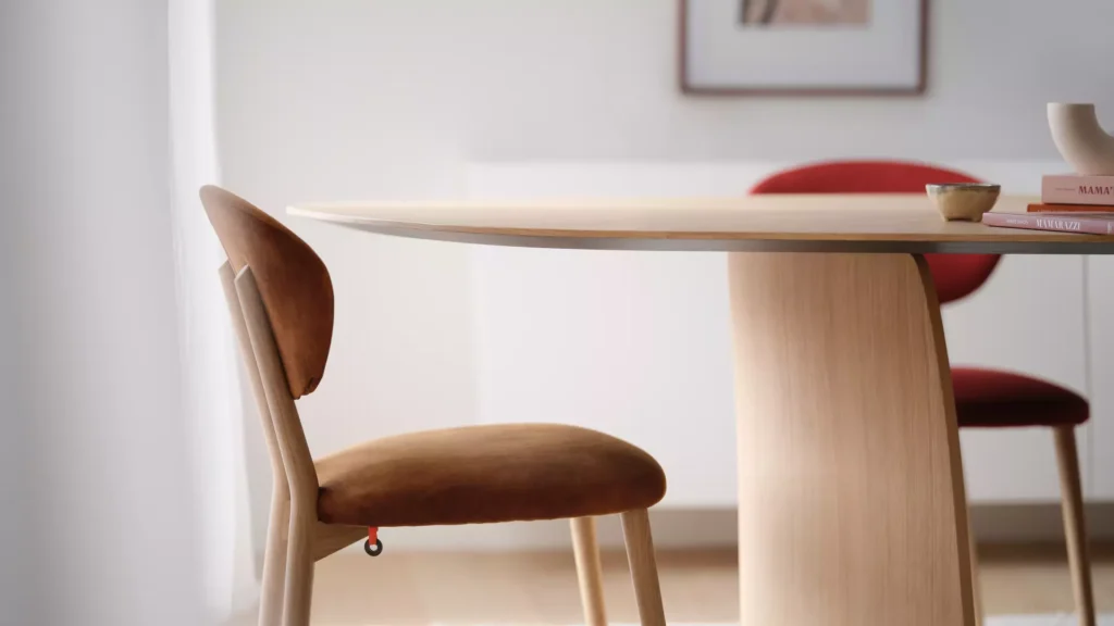 La table Dolmen Organic T0100 (120x300cm, chêne massif E) présente un bord incurvé et est associée à des chaises rembourrées brunes dans une pièce minimale, légèrement éclairée.