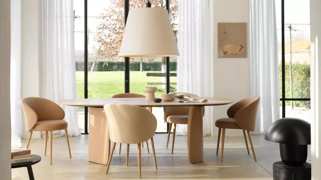 La table Dolmen Organic T0100 (120x300cm, chêne massif E), quatre chaises beiges, un décor minimal, de grandes fenêtres avec des rideaux blancs et une vue sur une pelouse ensoleillée constituent la base d'une salle à manger moderne.