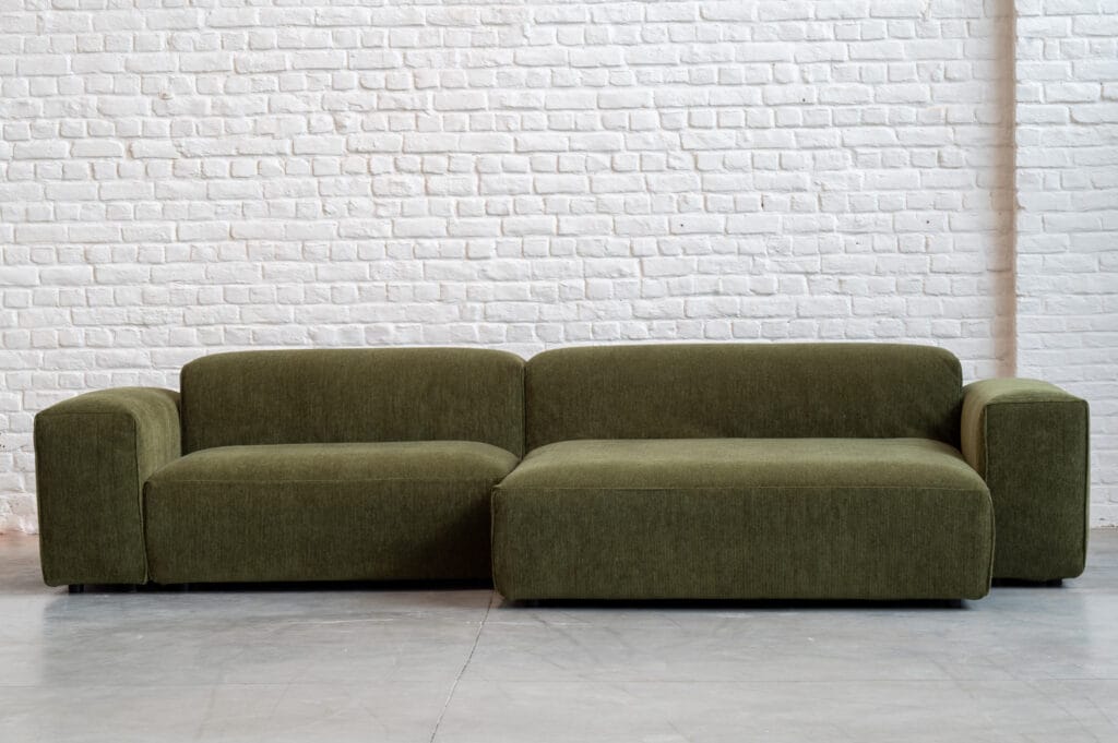 Un Canapé Edda, canapé sectionnel en tissu Moss devant un mur de briques blanches.