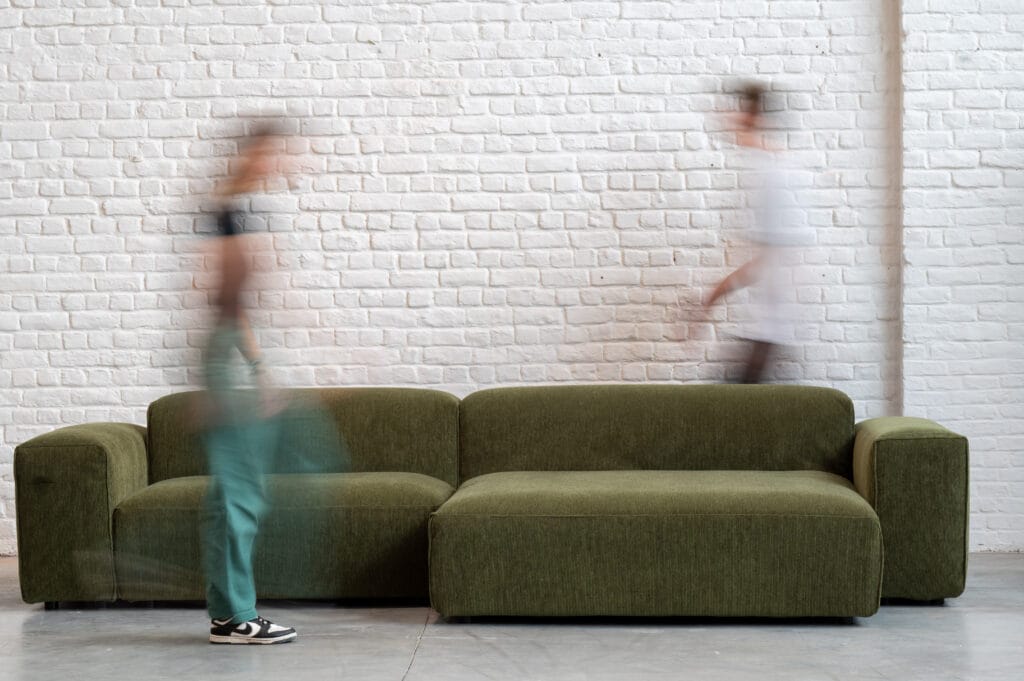 Deux personnes passant devant un Canapé Edda vert, en tissu Moss.