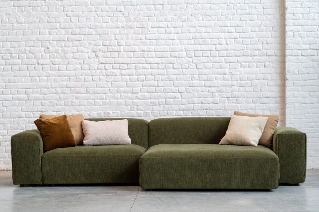 Un Canapé Edda, canapé en tissu vert mousse devant un mur de briques blanches.