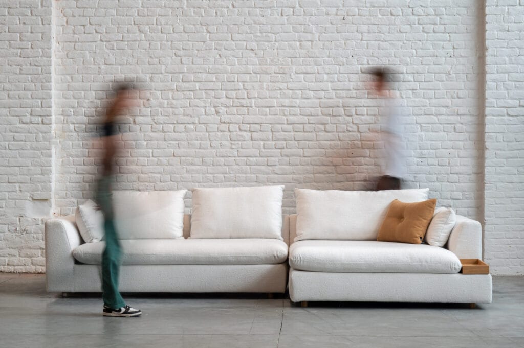 Deux personnes passant devant un Canapé Milou, tissu Heather dans une pièce au mur de briques.